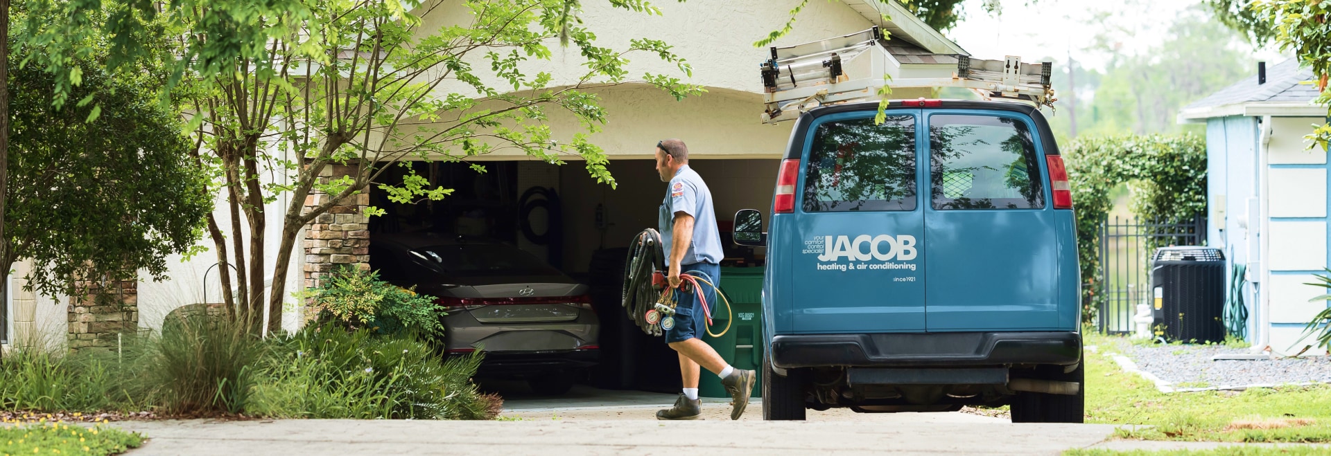 Jacob Heating & Air Conditioning technician walking away from company van with logo, while carrying equipment towards a house