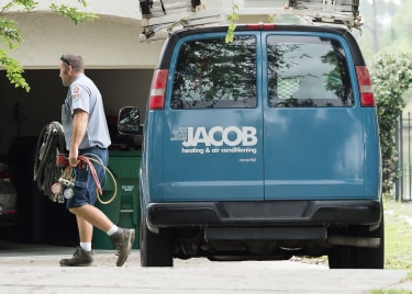 Jacob Heating & Air Conditioning technician walking away from company van with logo, while carrying equipment towards a house