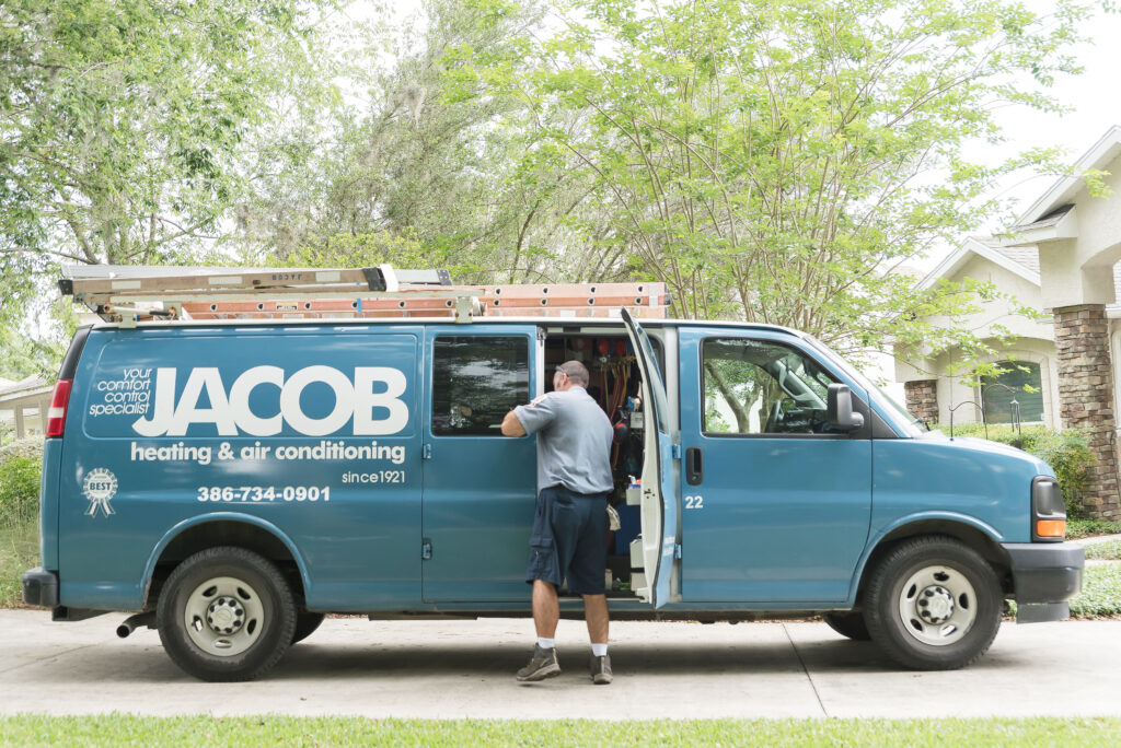 Technician opening doors of a blue Jacob service van parked in a home's driveway. Green trees in background.