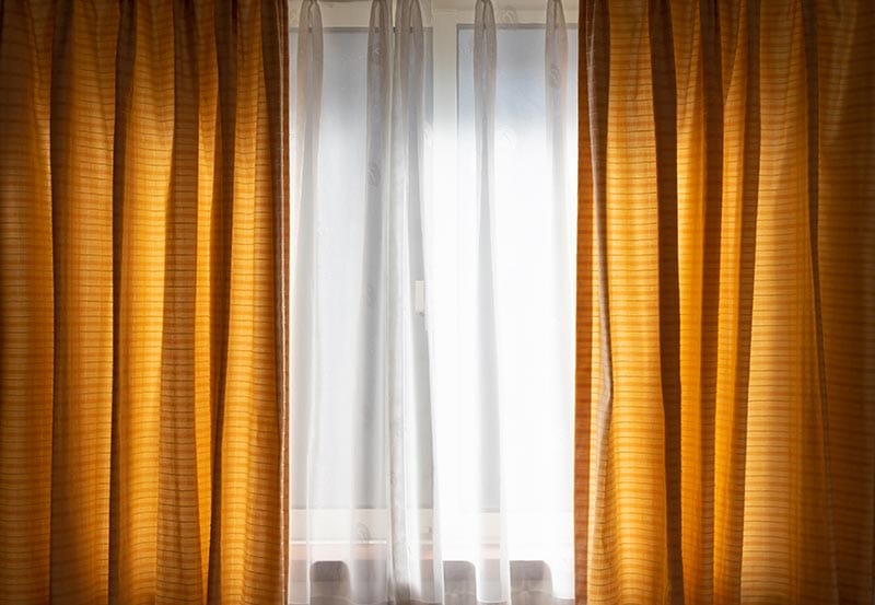 A window framed by heavy mustard-yellow drapes, with translucent white sheer curtains drawn across the center letting soft daylight filter through.