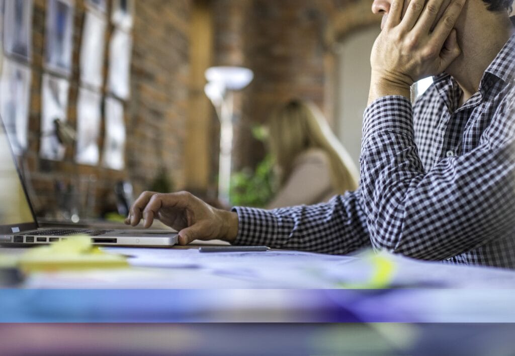Unrecognizable white male sitting behind a desk in modern office with one hand propped up by face and other using mobile device.
