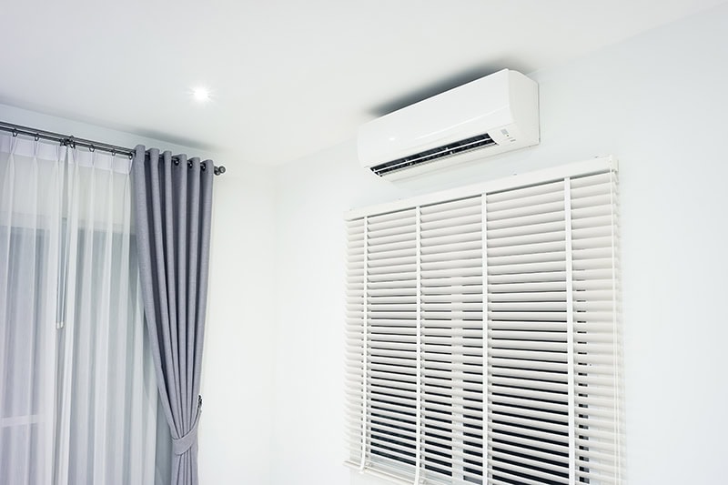 A white ductless mini-split air conditioner mounted high on a white wall above a window with white horizontal blinds, set in a modern room with grey curtains.