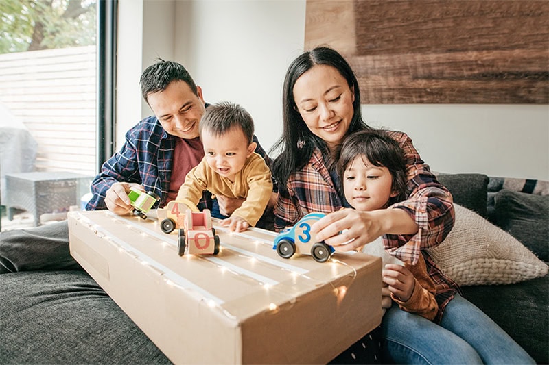 A family of four sitting together indoors on a couch, smiling as the parents play with toy wooden cars on a cardboard box track with their two young children.